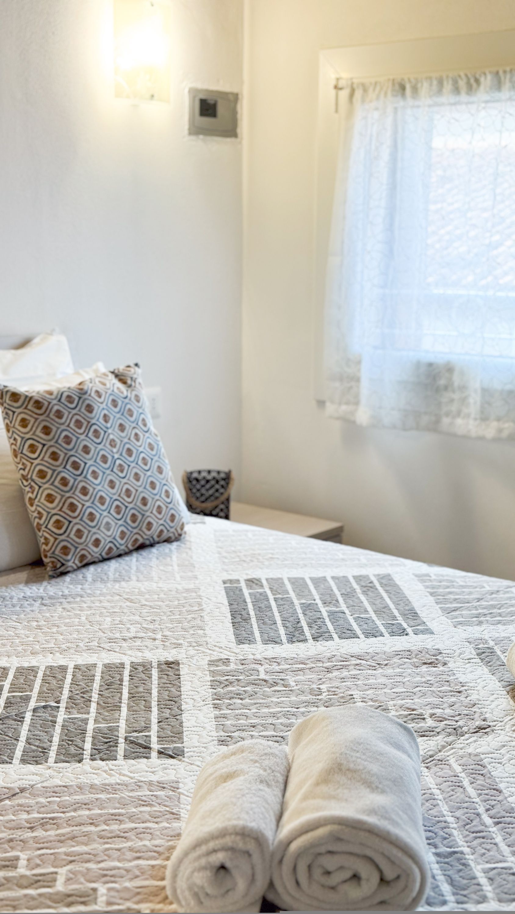 Una cama bien hecha con toallas enrolladas, un cojín estampado y una mesita de noche pequeña.