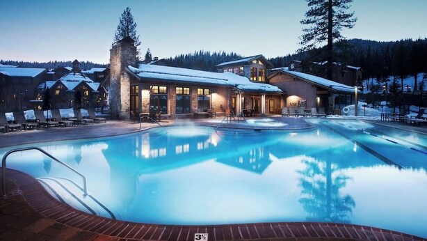 Un resort con una amplia piscina exterior, cabañas de madera y montañas al fondo.