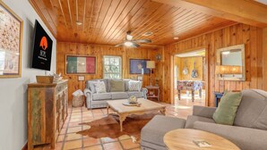 A cozy cabin interior with wooden walls and ceiling, a sofa, a coffee table, and a dining area visible through an open door. - Lawlis Lodge I Families, Star Gazing (Wimberley)