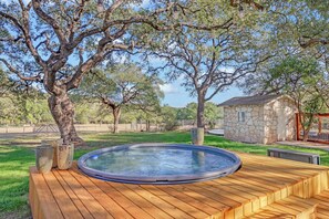 Een houten terras met een jacuzzi, omgeven door bomen en een stenen gebouw.