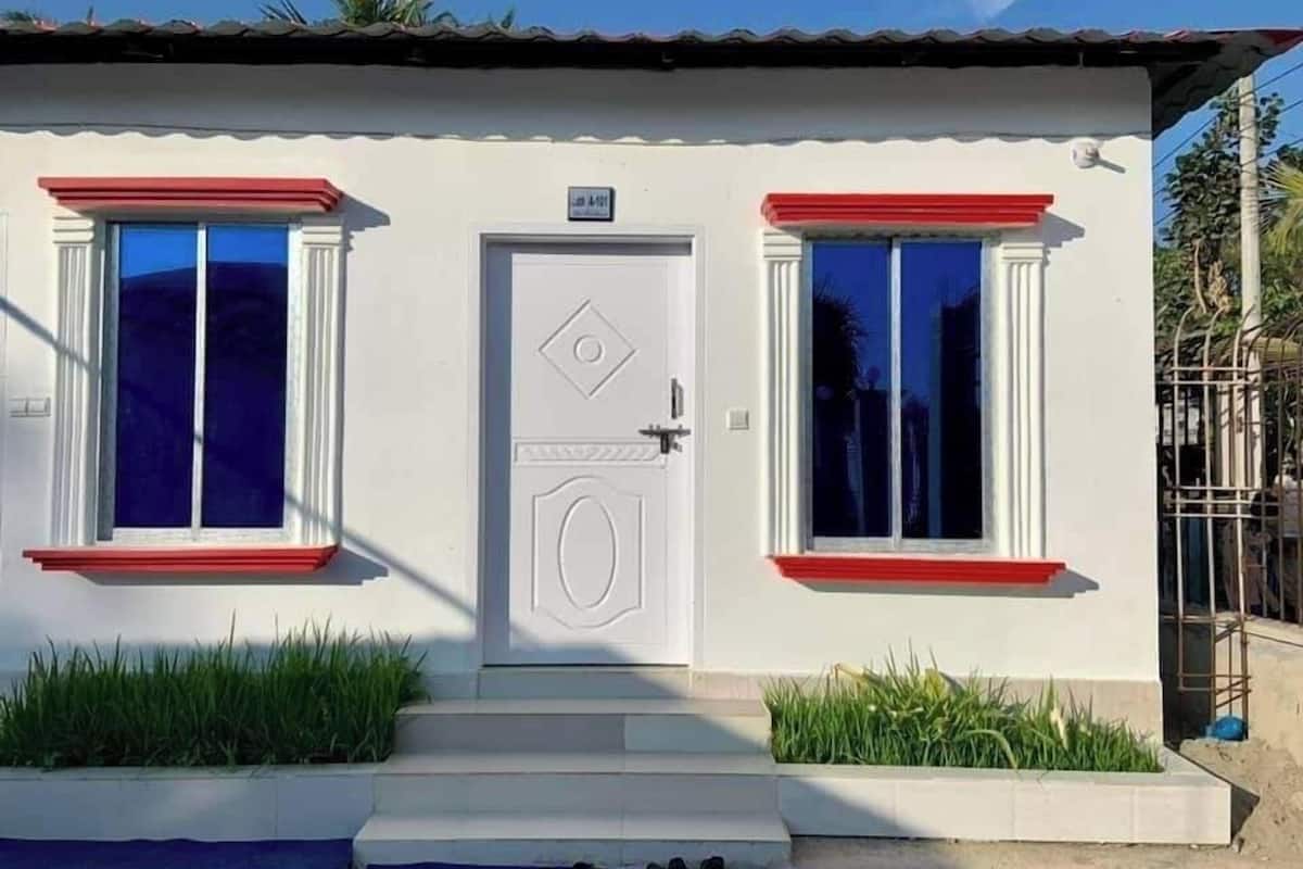 Un pequeño edificio de una sola planta con fachada blanca, puerta enmarcada en rojo y dos ventanas con marcos rojos.