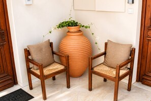 Dos sillas de madera con asientos de mimbre y cojines, un gran jarro de terracota con una planta, y una puerta de madera con felpudo.