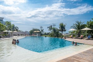 Une piscine entourée de chaises longues et de parasols, bordée de palmiers et surplombée d’un ciel dégagé.