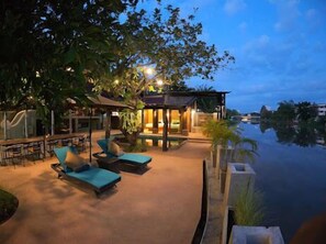 A poolside area with lounge chairs and a covered seating area, surrounded by trees and a view of the water.