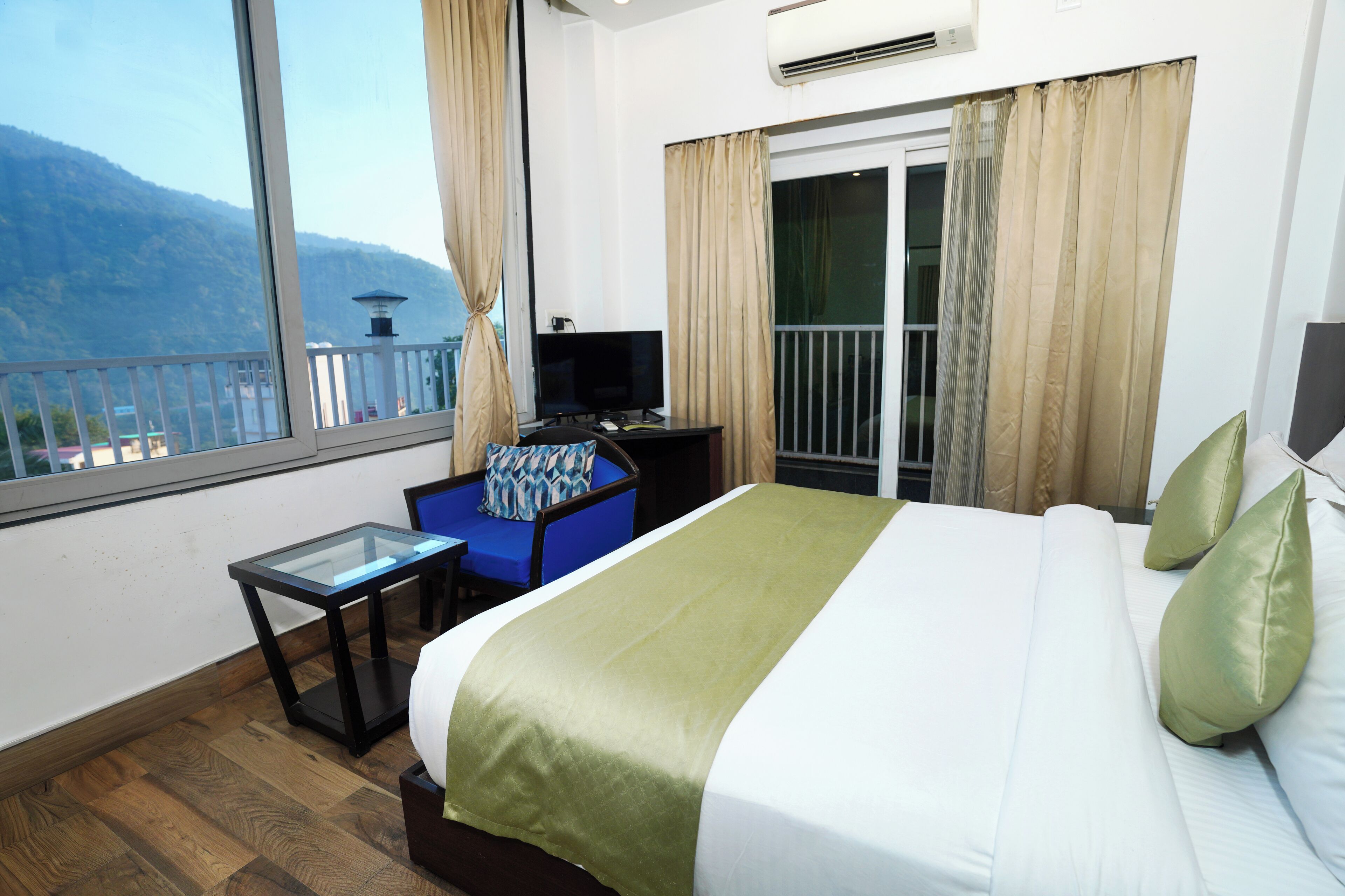 A hotel room with a large bed, a desk with a computer, a TV, and a view of mountains through a large window.