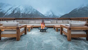 Un área de estar al aire libre con suelo de piedra, mobiliario de madera y una chimenea central.