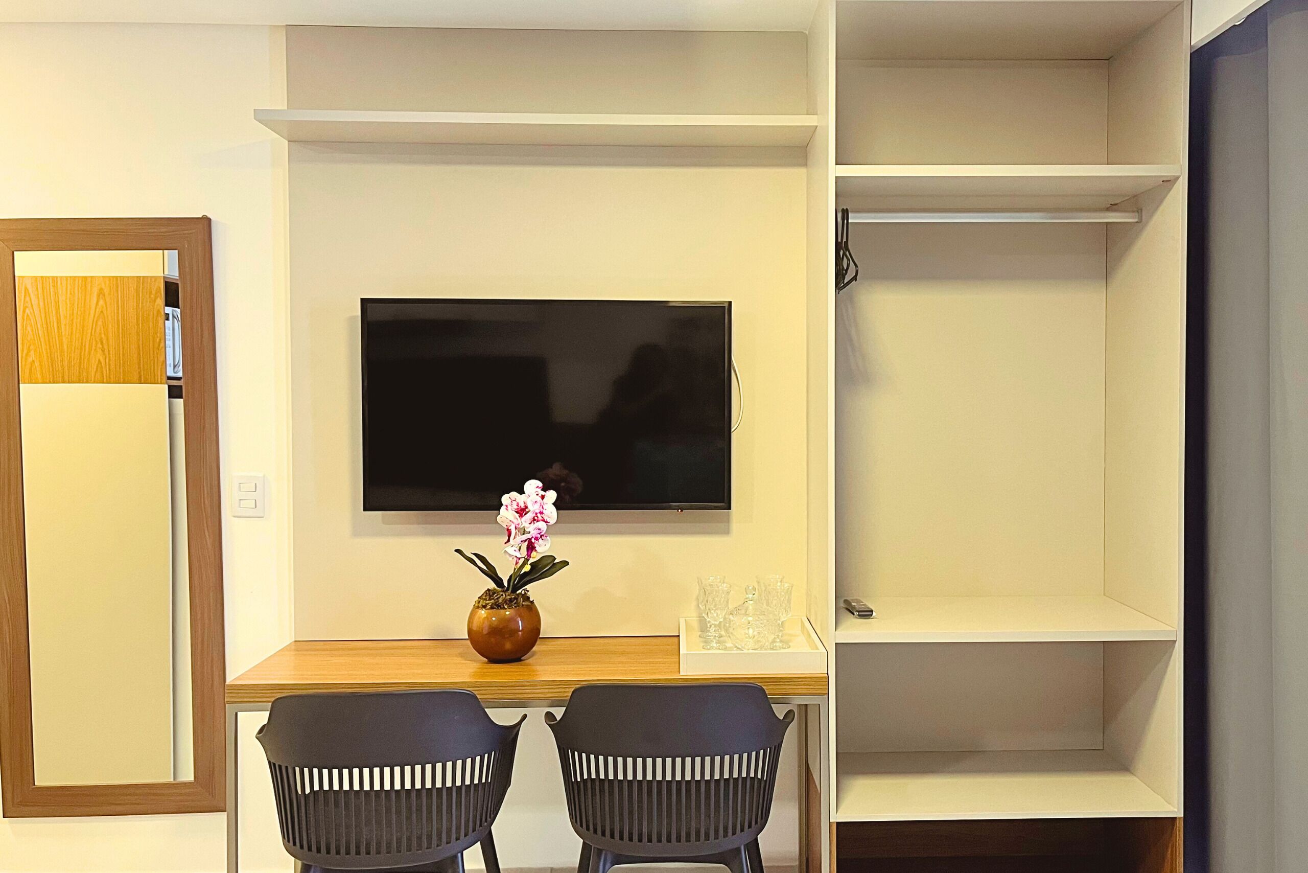Une pièce avec un téléviseur fixé au mur, un bureau en bois avec deux chaises, un vase de fleurs et une étagère avec une télécommande.