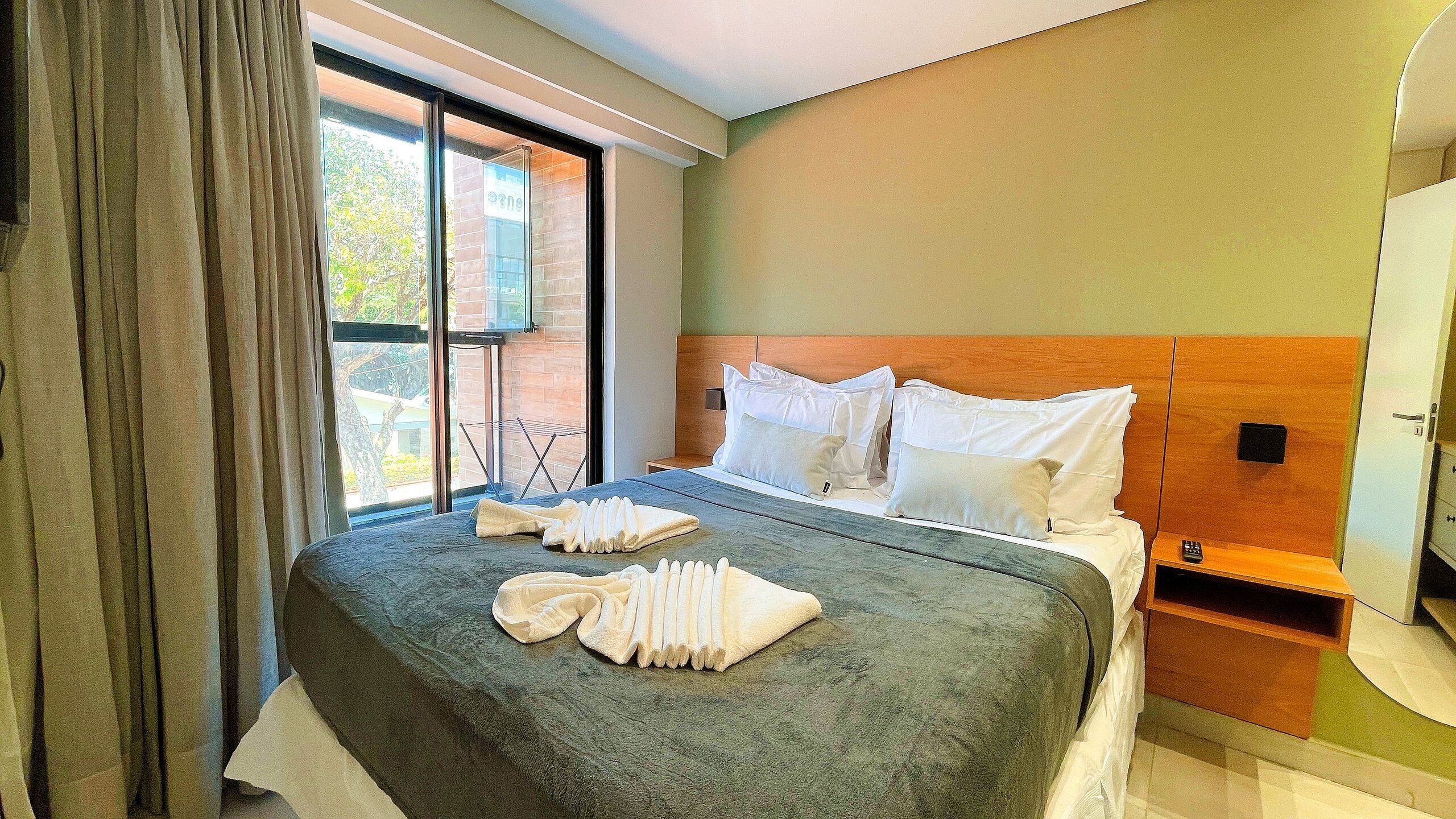 A hotel room with a large bed, a wooden headboard, a bedside table, and a window with curtains.