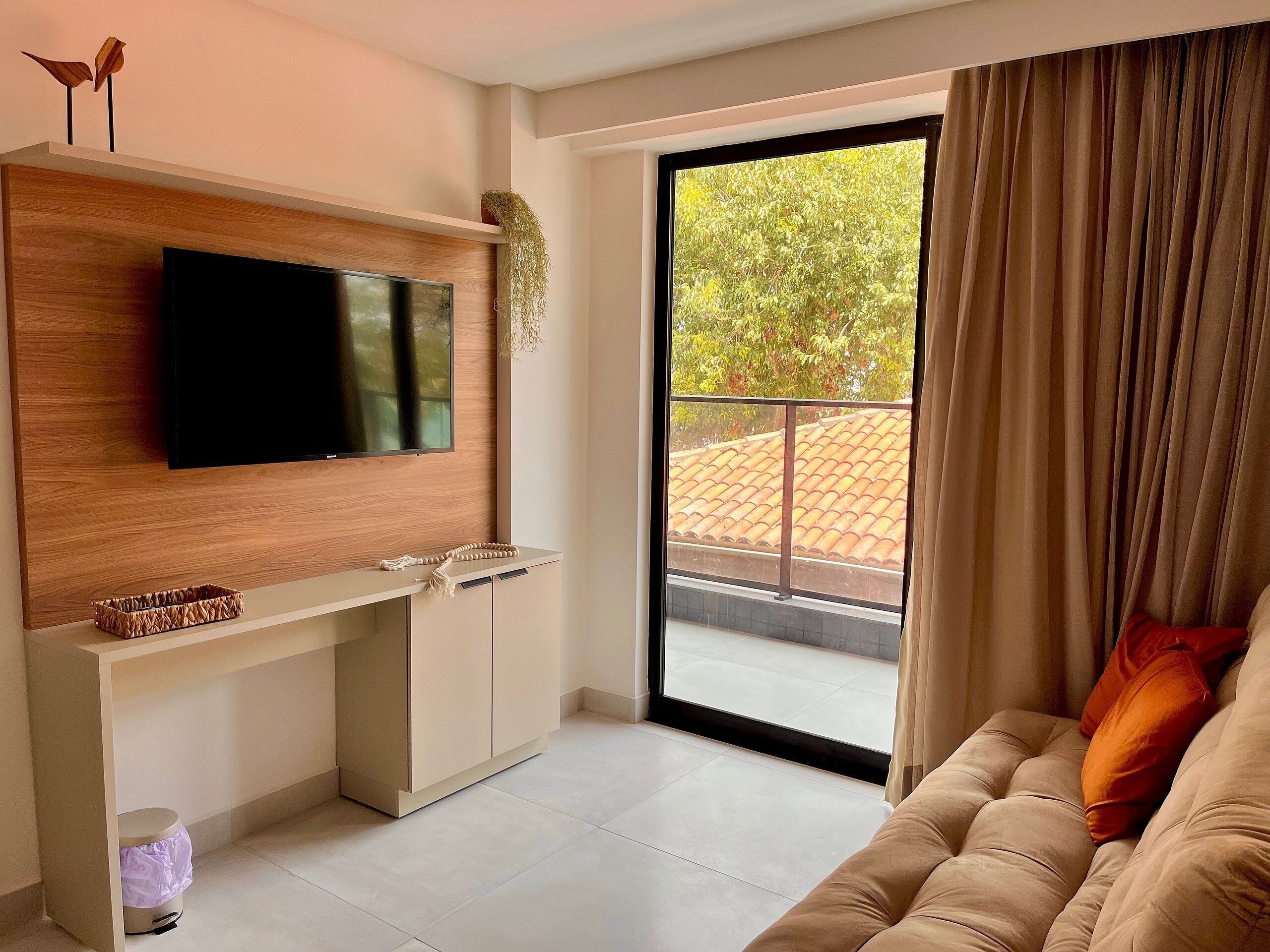 A modern living room with a flat-screen TV mounted on a wooden wall, a sliding glass door leading to an outdoor area, and a beige sofa with a cushion.
