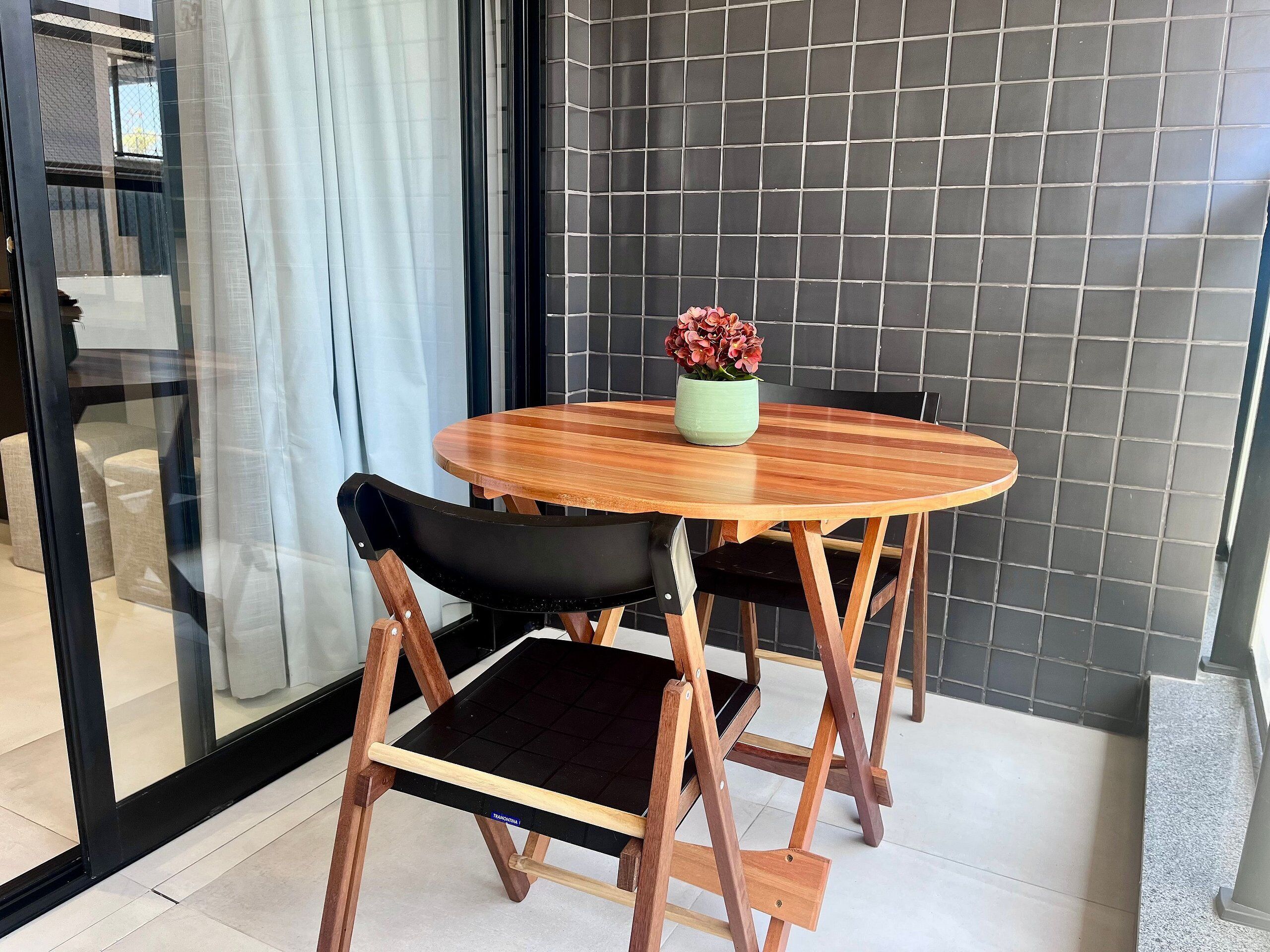 A round wooden table with a black cushioned chair, a potted plant, and a tiled wall in the background.