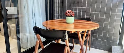 A round wooden table with a black cushioned chair, a potted plant, and a tiled wall in the background.