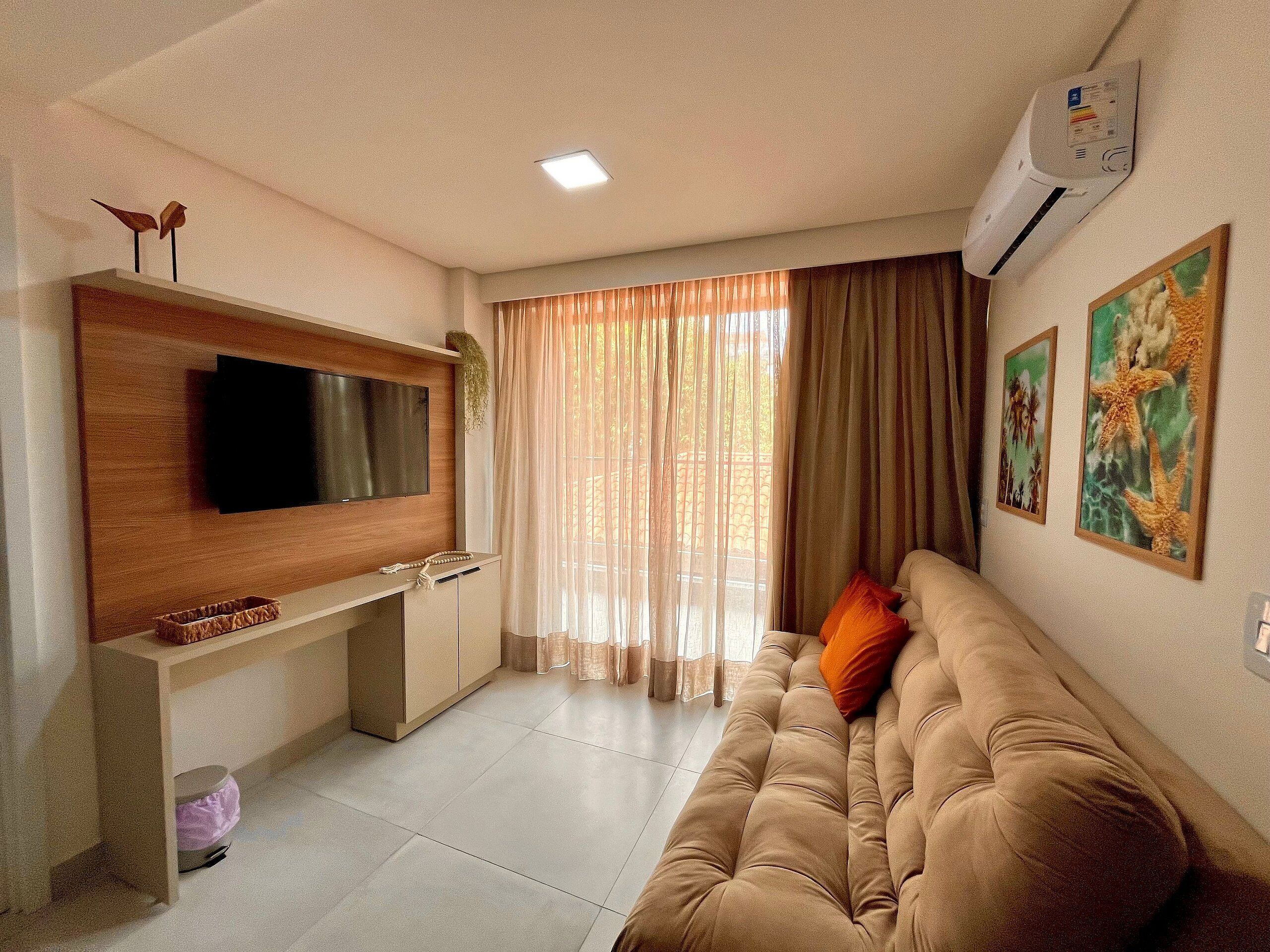 A living room with a flat-screen TV mounted on a wooden console, a beige sofa with orange cushions, and a large window with sheer curtains.