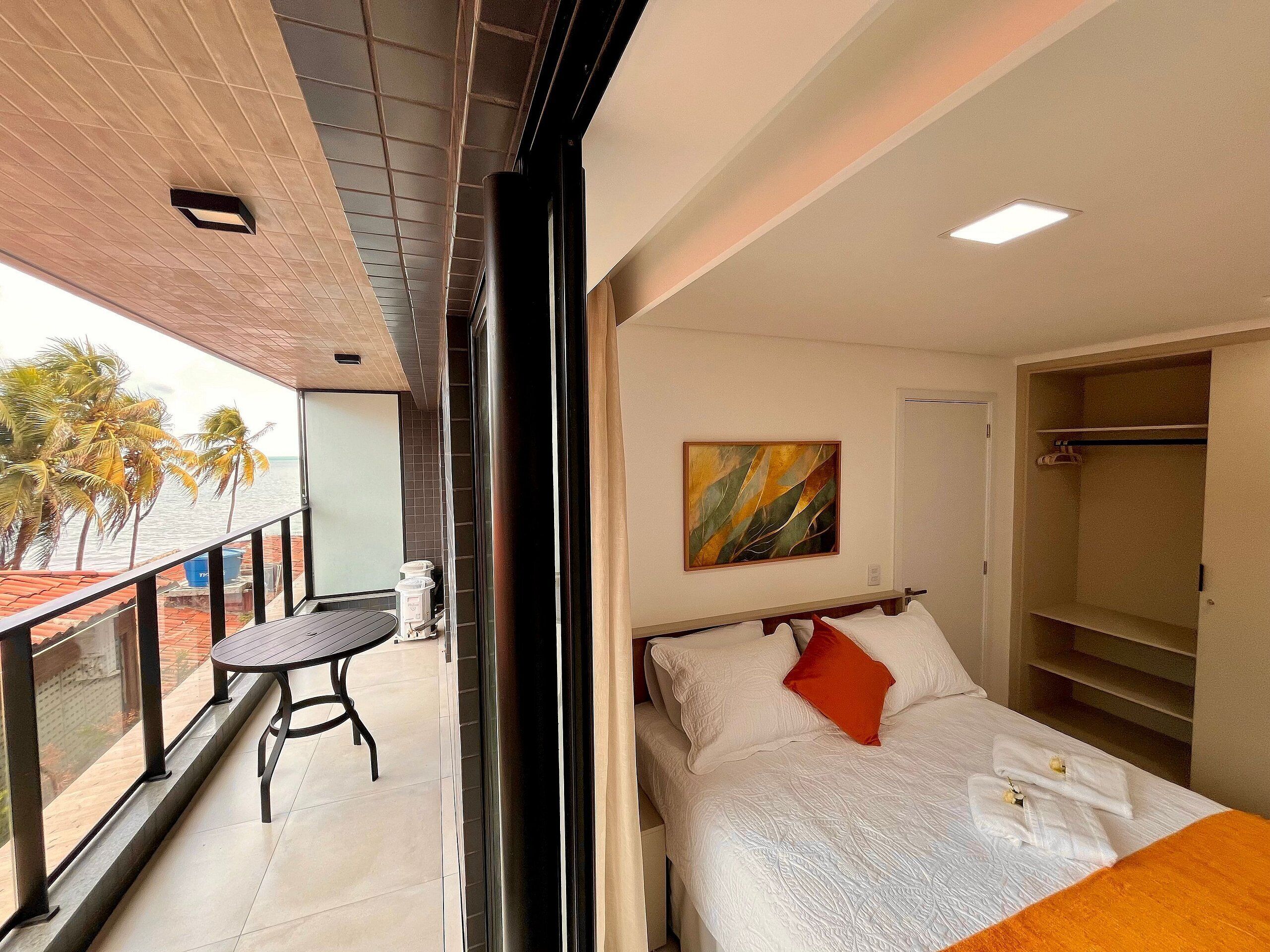A hotel room with a balcony, a bed with white bedding and an orange pillow, a painting on the wall, and a wardrobe.