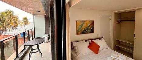 A hotel room with a balcony, a bed with white bedding and an orange pillow, a painting on the wall, and a wardrobe.