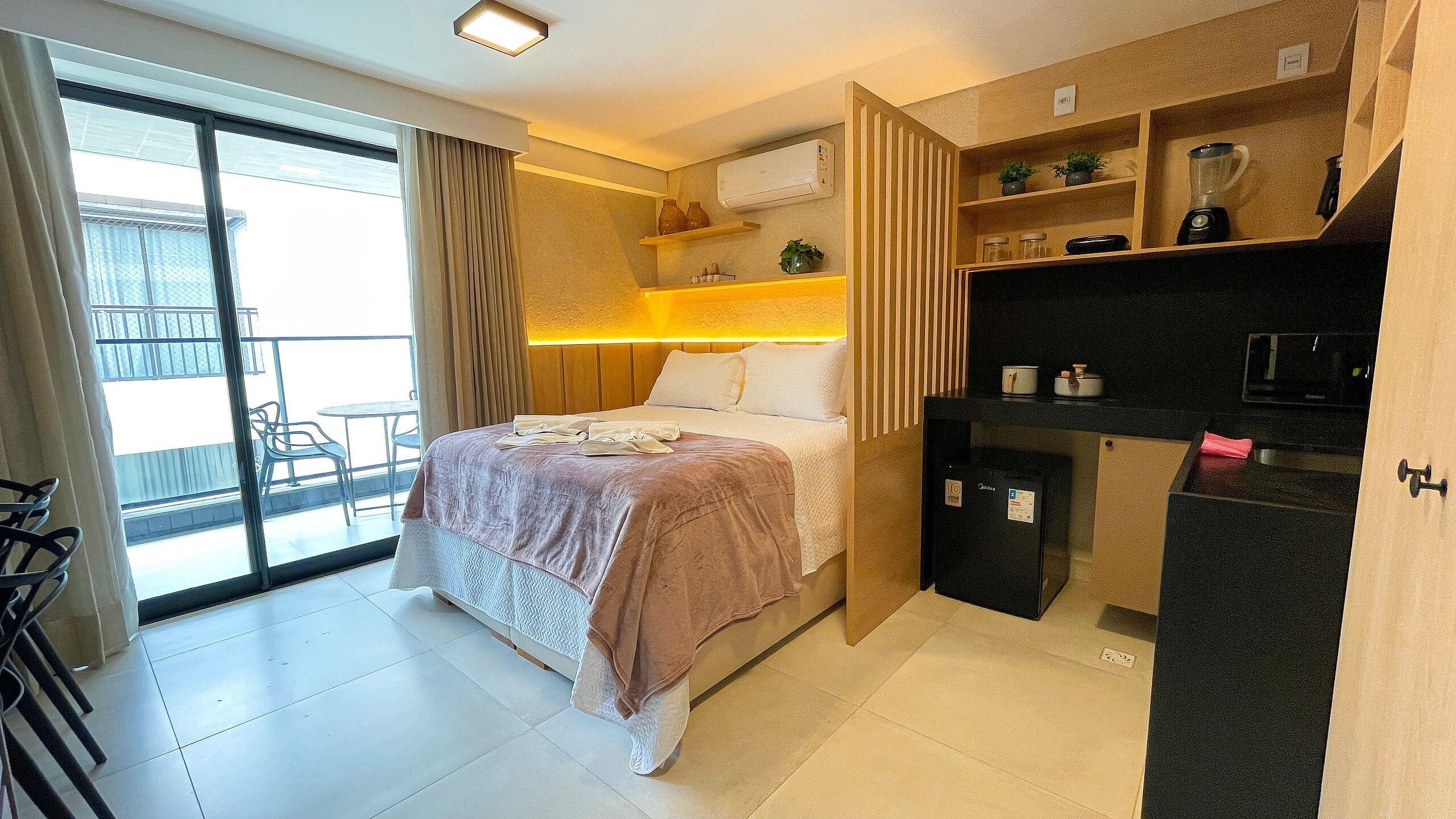 A hotel room with a bed, a desk, a refrigerator, and a view of the outside balcony.