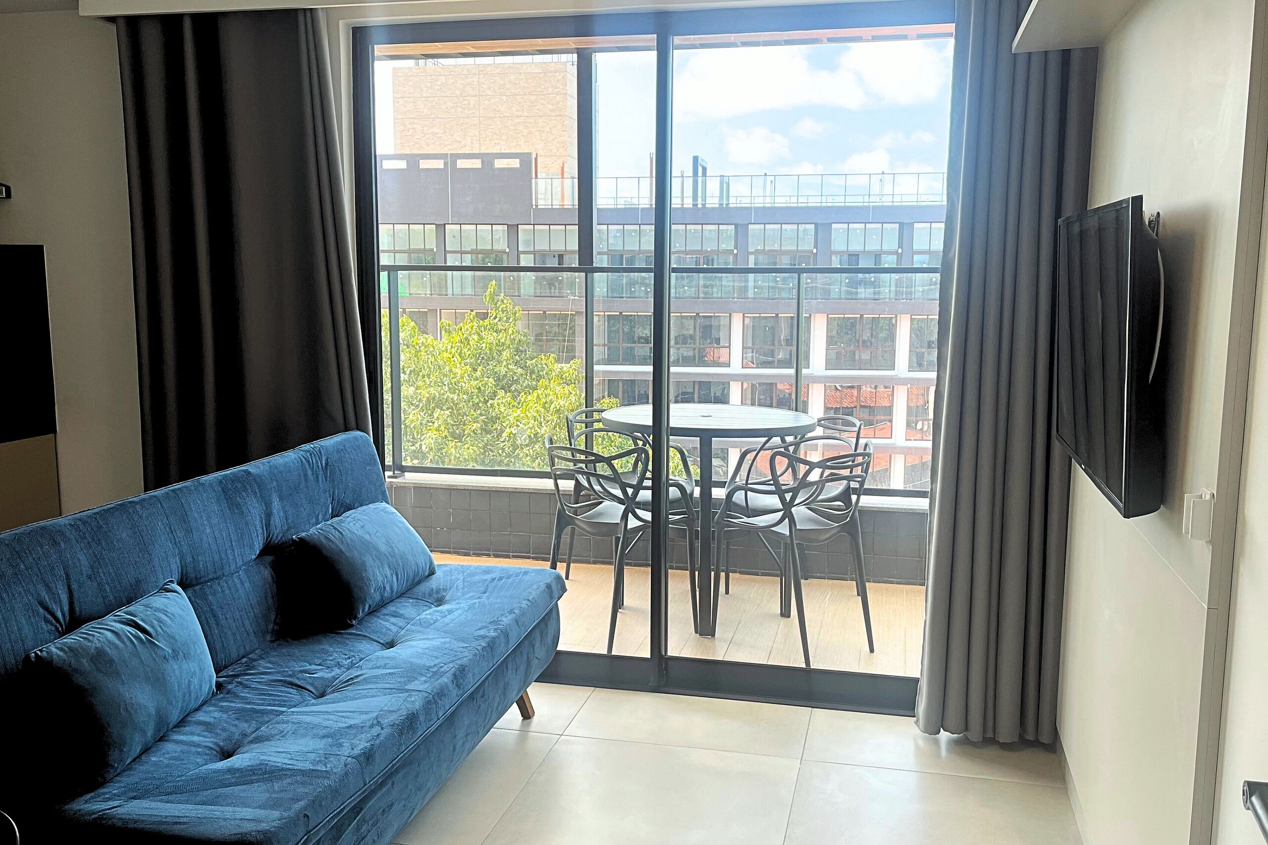 A modern living room with a blue sofa, a round table with chairs, and a large window offering a view of buildings and greenery.