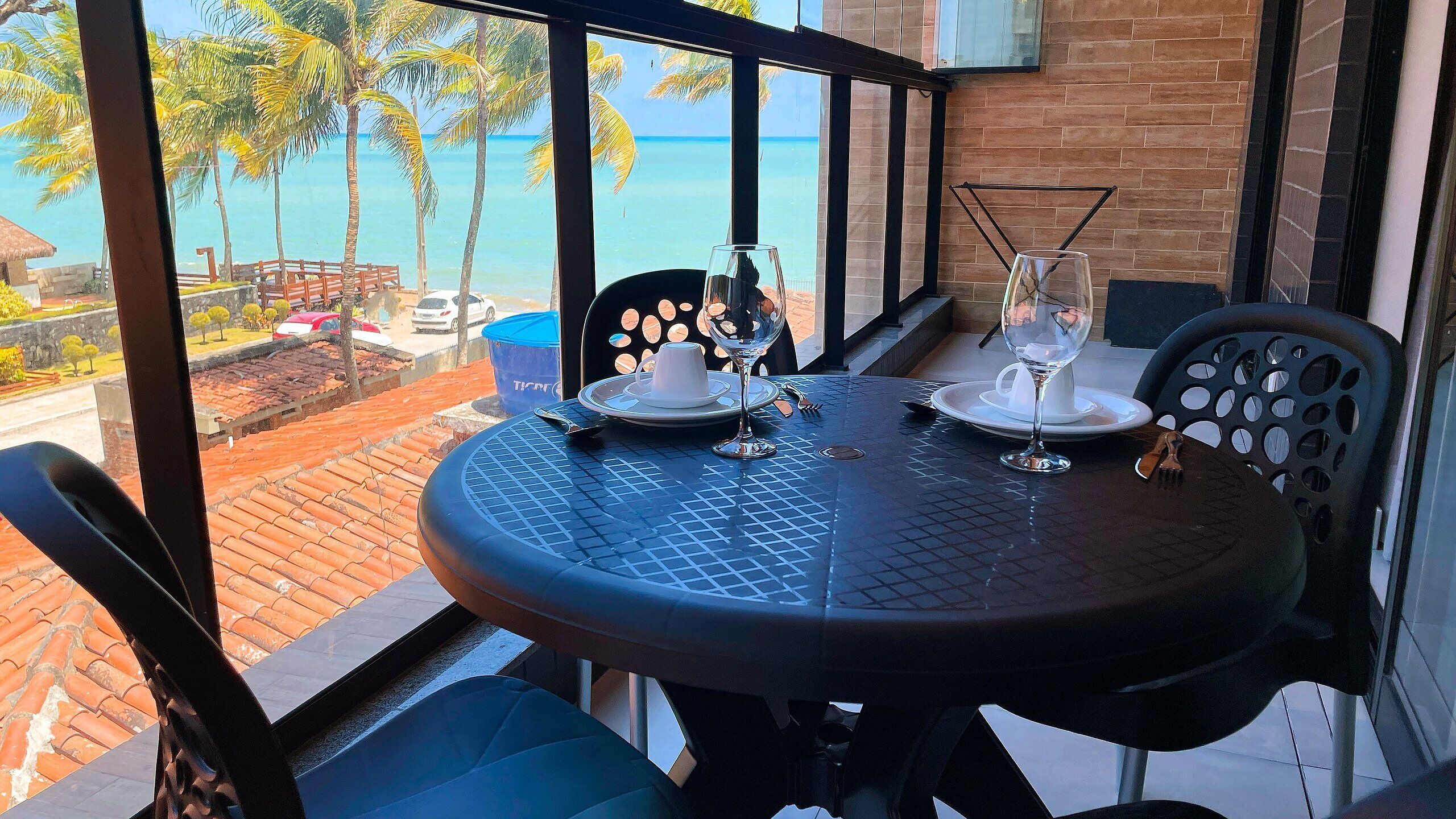 Uma mesa de jantar com cadeiras, um copo de água e uma taça de vinho, com vista para palmeiras e o oceano.