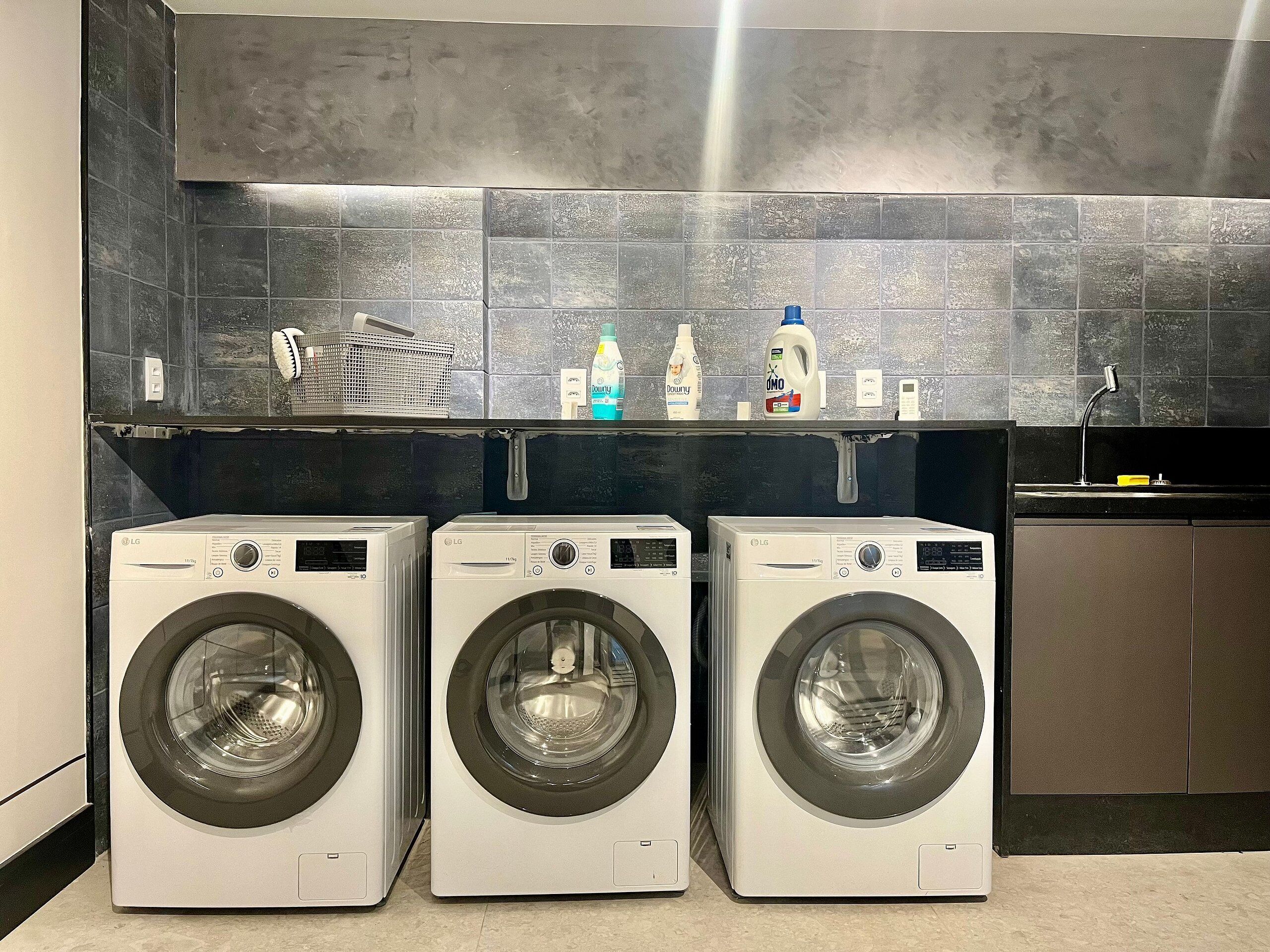 A laundry area with three LG washing machines, a shelf with cleaning products, and a black tiled backsplash.