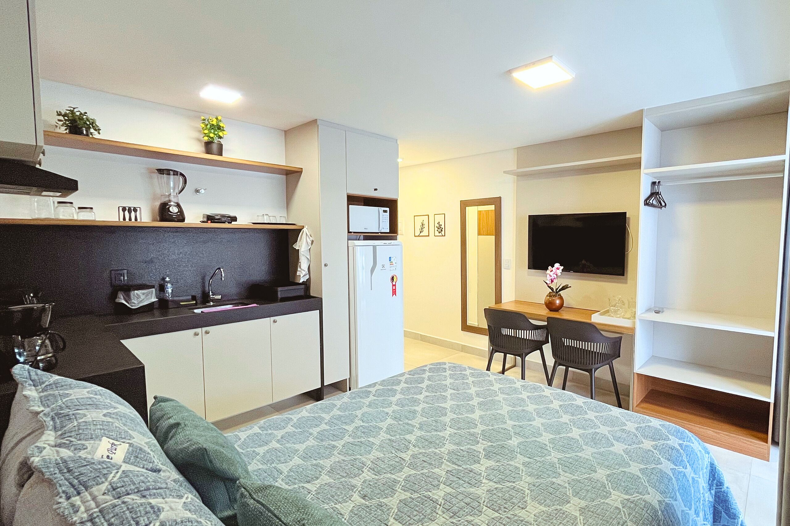 Ein kompaktes Hotelzimmer mit Küchenzeile, einem Bett mit gemusterten Textilien, einer Essecke mit Stühlen und einem an der Wand montierten Fernseher.