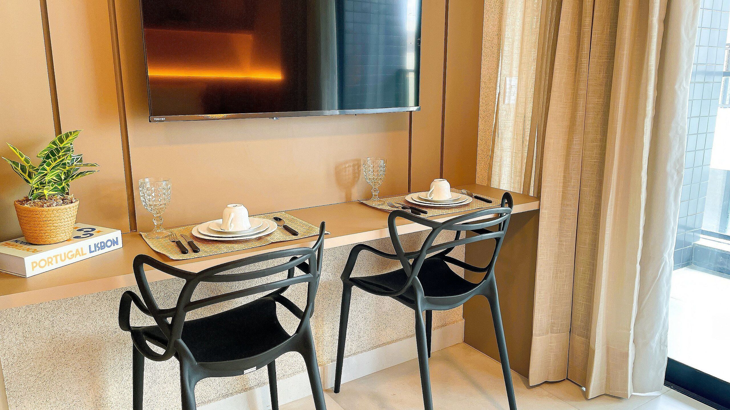 A dining area with two chairs, a table, a TV mounted on the wall, a potted plant, and a book titled "Portugal Lisbon".