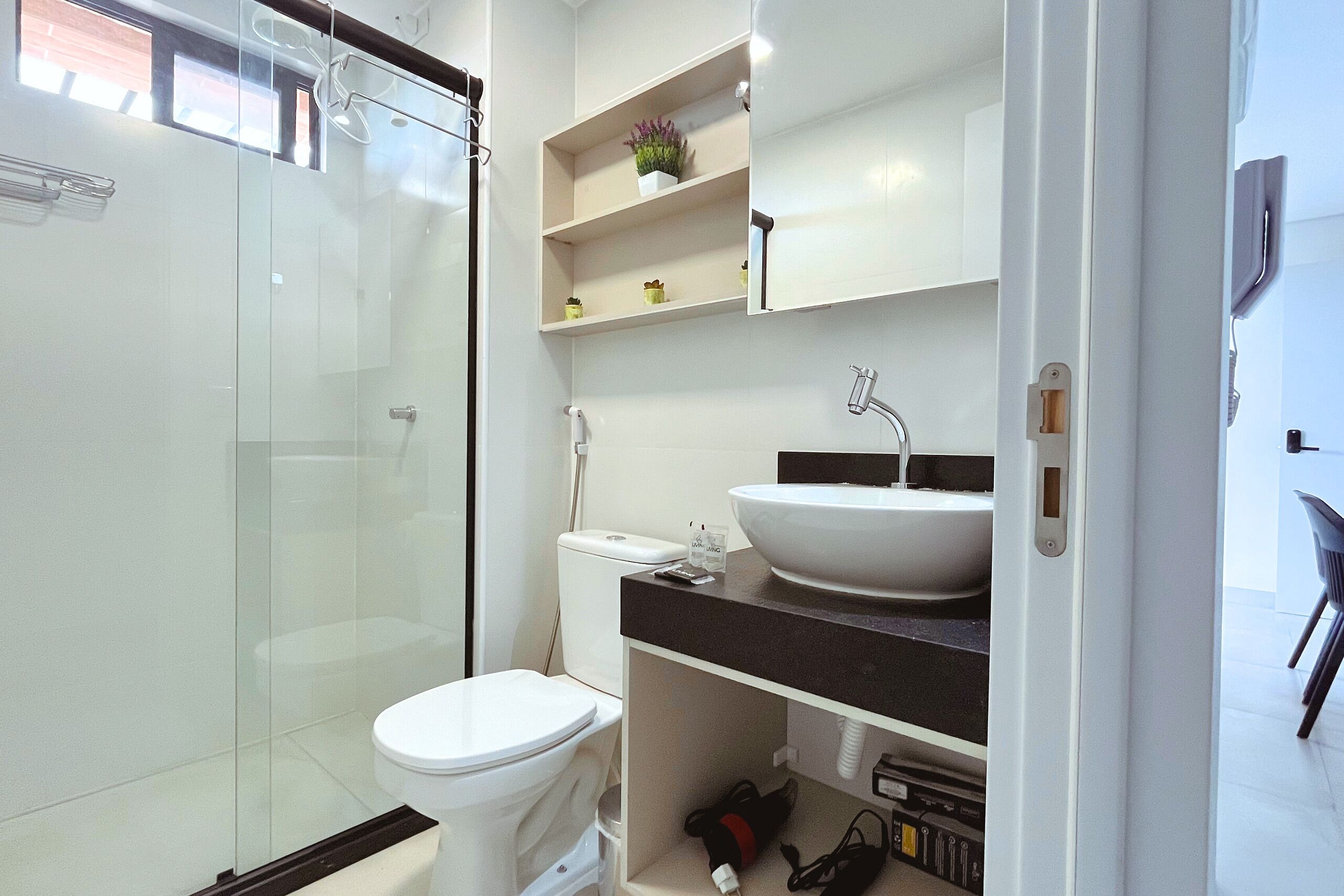 A modern bathroom with a glass shower enclosure, a white toilet, a white sink with a black countertop, and a shelf with decorative items.