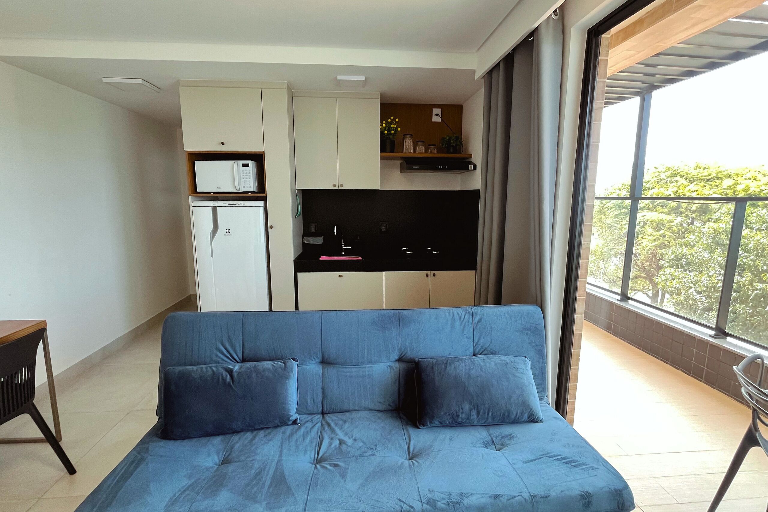 Een moderne woonkamer met een blauwe bank, een klein keukenhoekje met een magnetron, en een balkon met uitzicht op bomen.