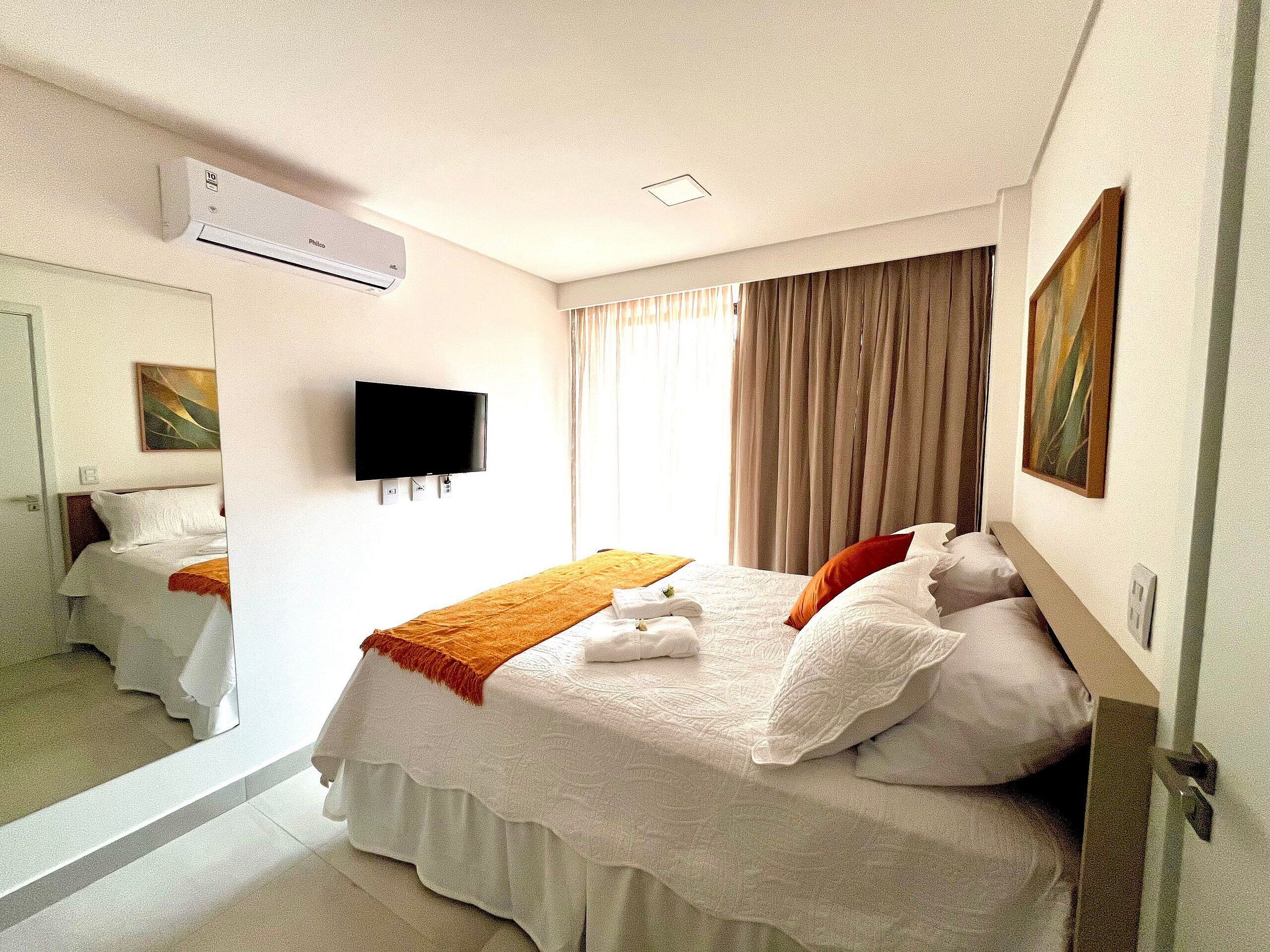 A hotel room with a bed, a flat-screen TV, an air conditioning unit, a window with curtains, and a painting on the wall.