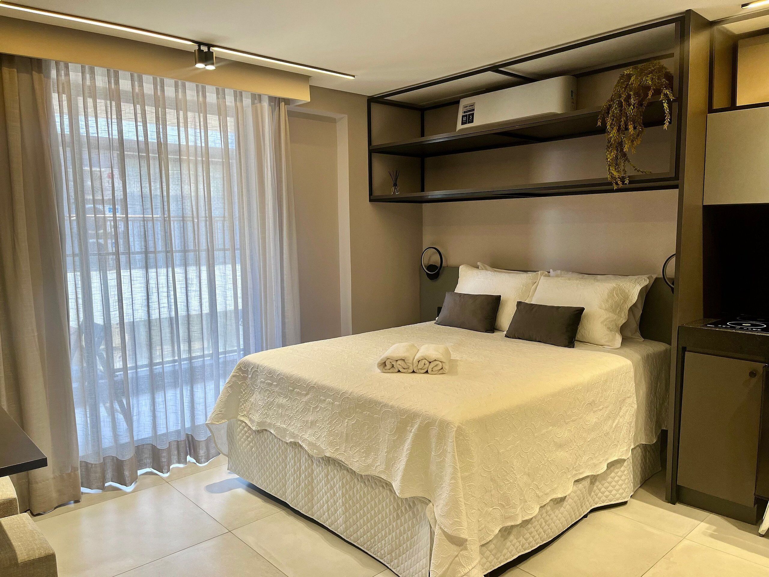A modern hotel room with a neatly made bed, a large window with sheer curtains, and built-in shelves above the bed.