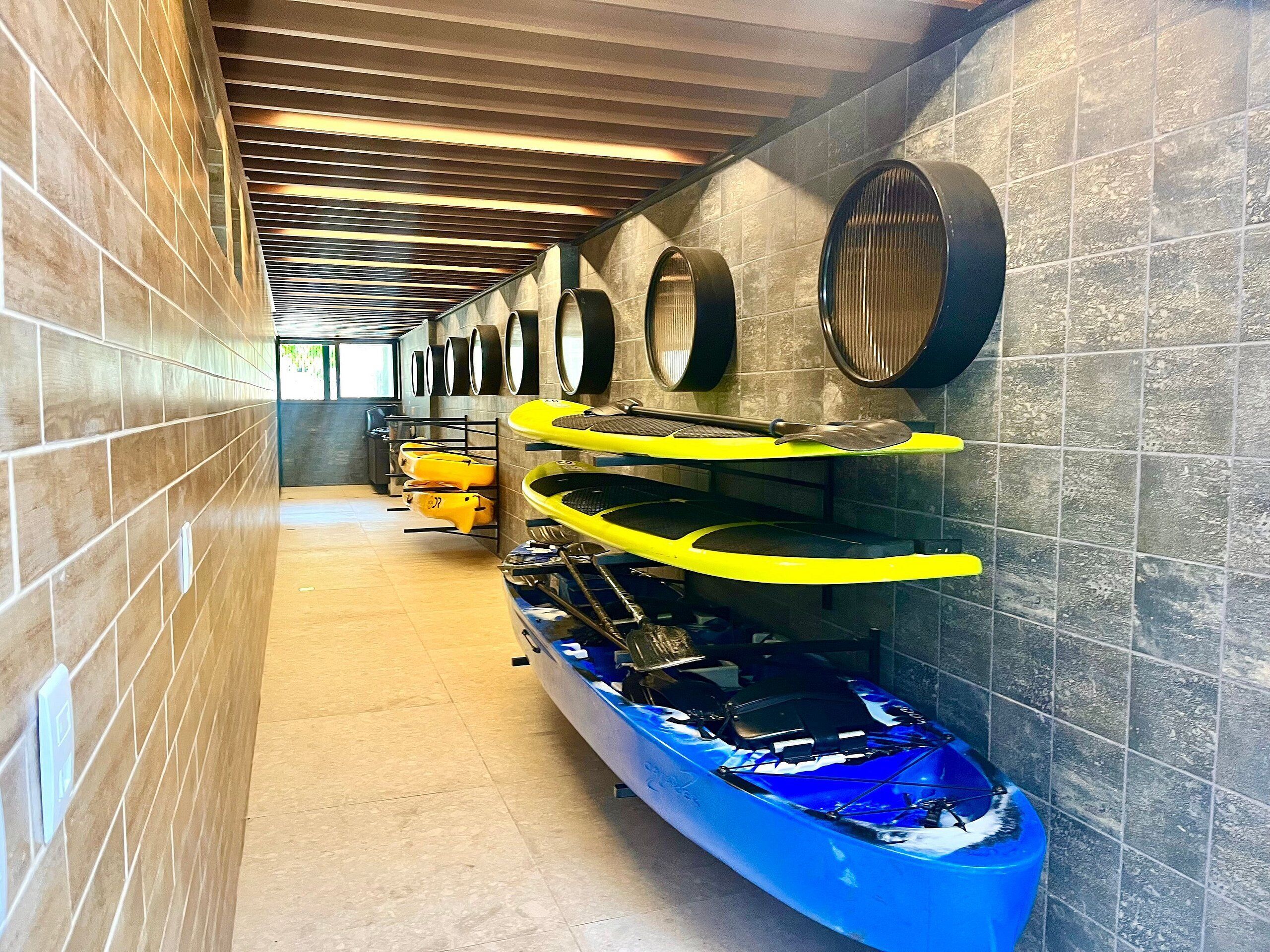 Una serie di kayak e tavole da paddleboard in un'area coperta con pareti in piastrelle e soffitto in legno.