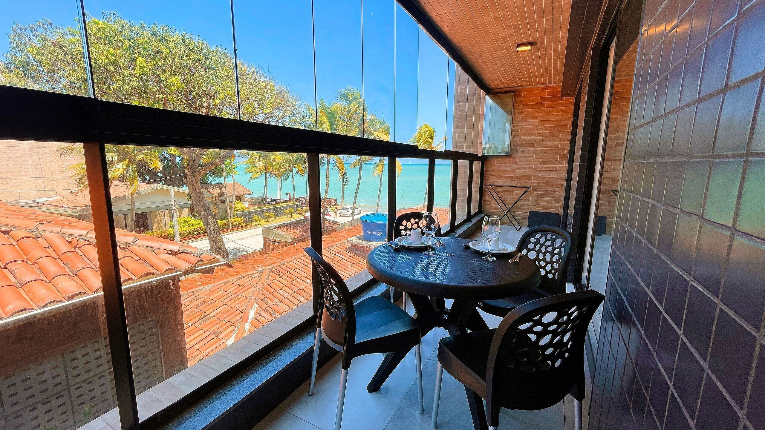 A balcony with a table and chairs, overlooking a coastal area with palm trees and a clear blue sky.