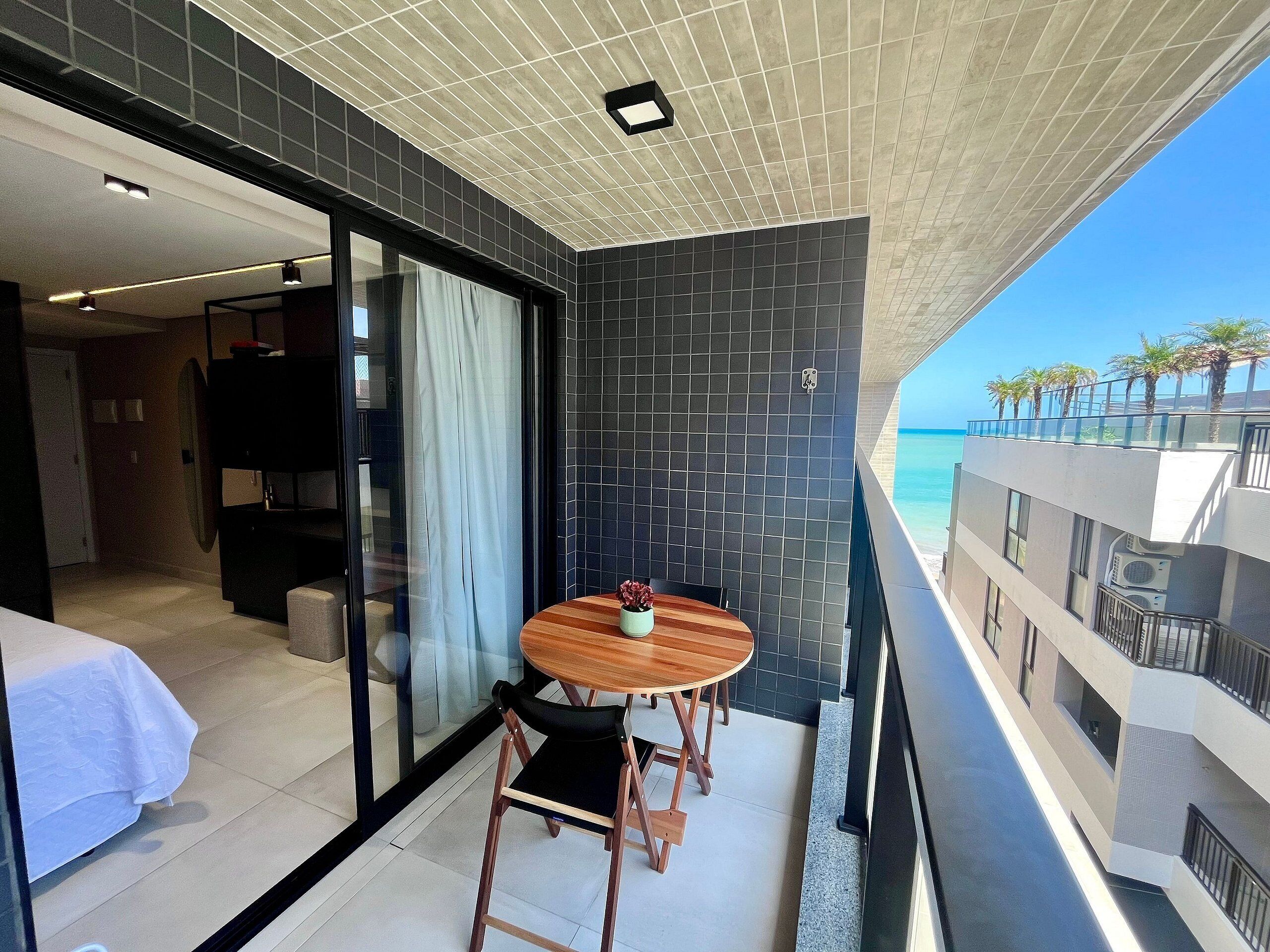 Habitación de hotel moderna con balcón y vistas a la playa. Incluye una cama grande, una pequeña mesa con sillas y vista al océano.