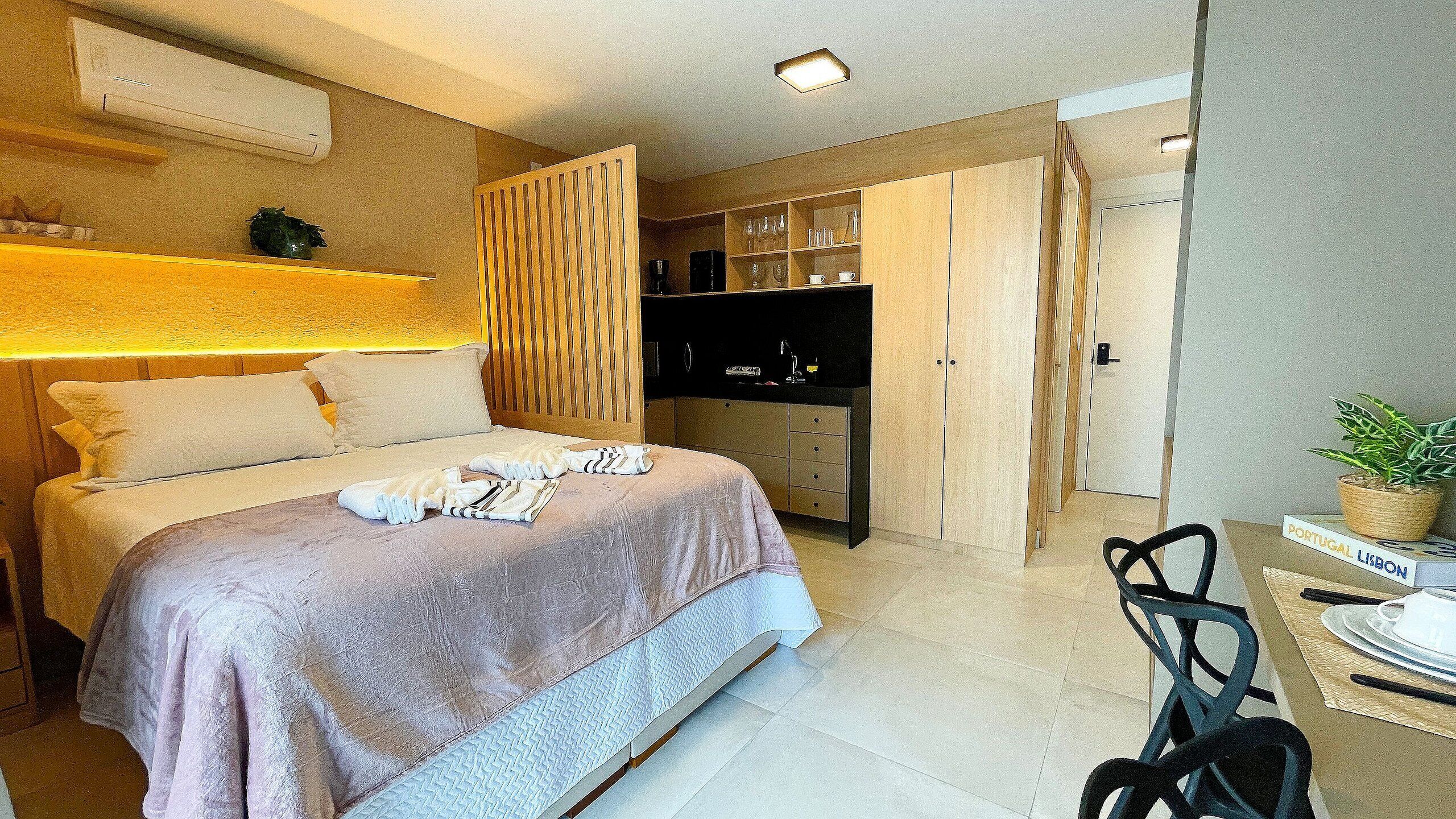 A hotel room with a bed, a desk with a chair, a wardrobe, and a small kitchen area.