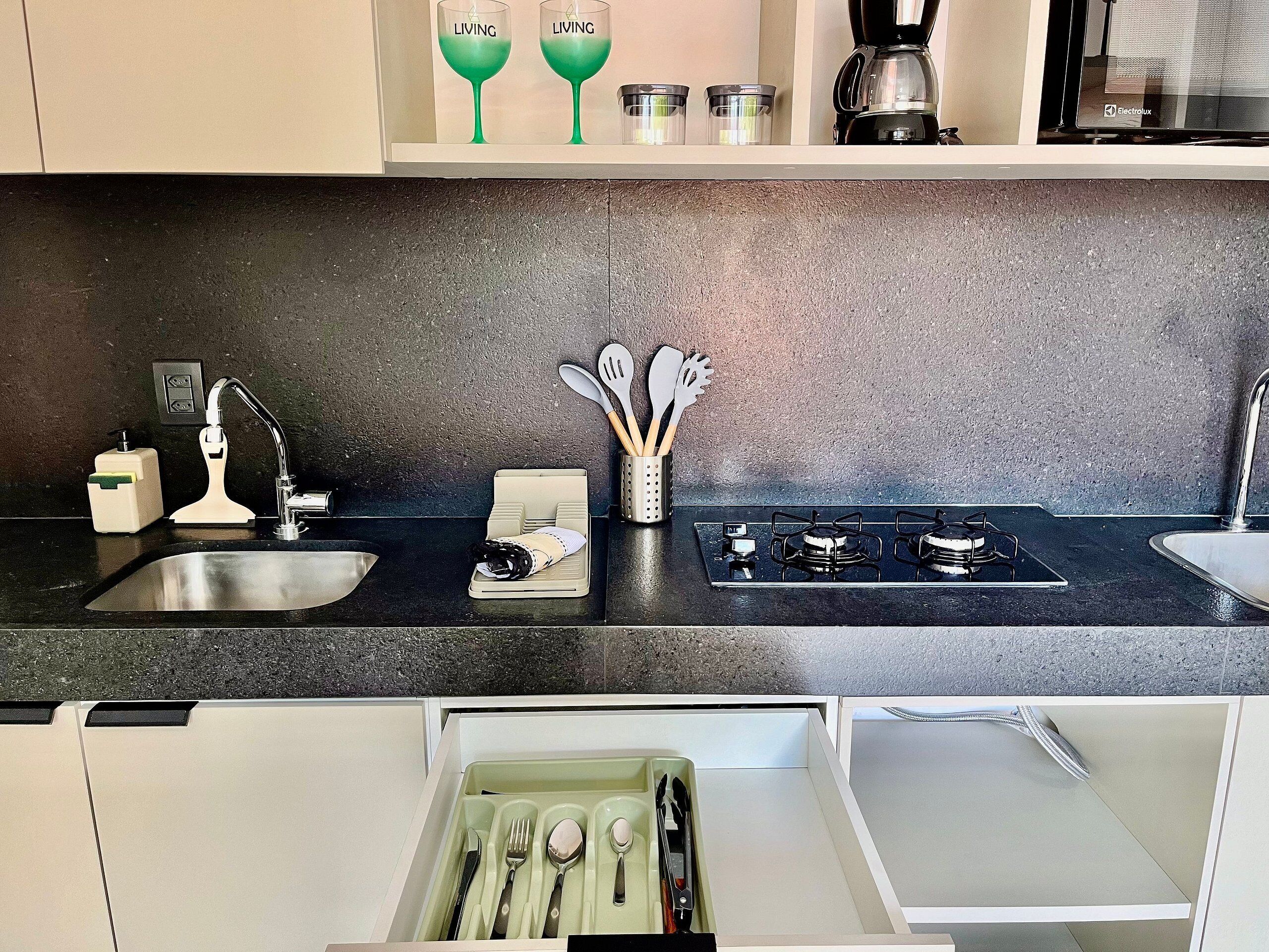 A kitchen with a black countertop, stainless steel sink, and a gas stove. There are two wine glasses on a shelf above the stove, and a coffee maker on the right.