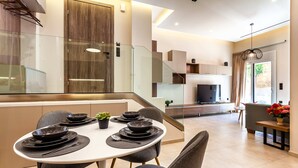A modern dining area with a white table set for four, surrounded by dark chairs. A glass partition separates the dining area from the kitchen. - Kalamata Gem Retreats, Hidden In the City Center (Kalamata)