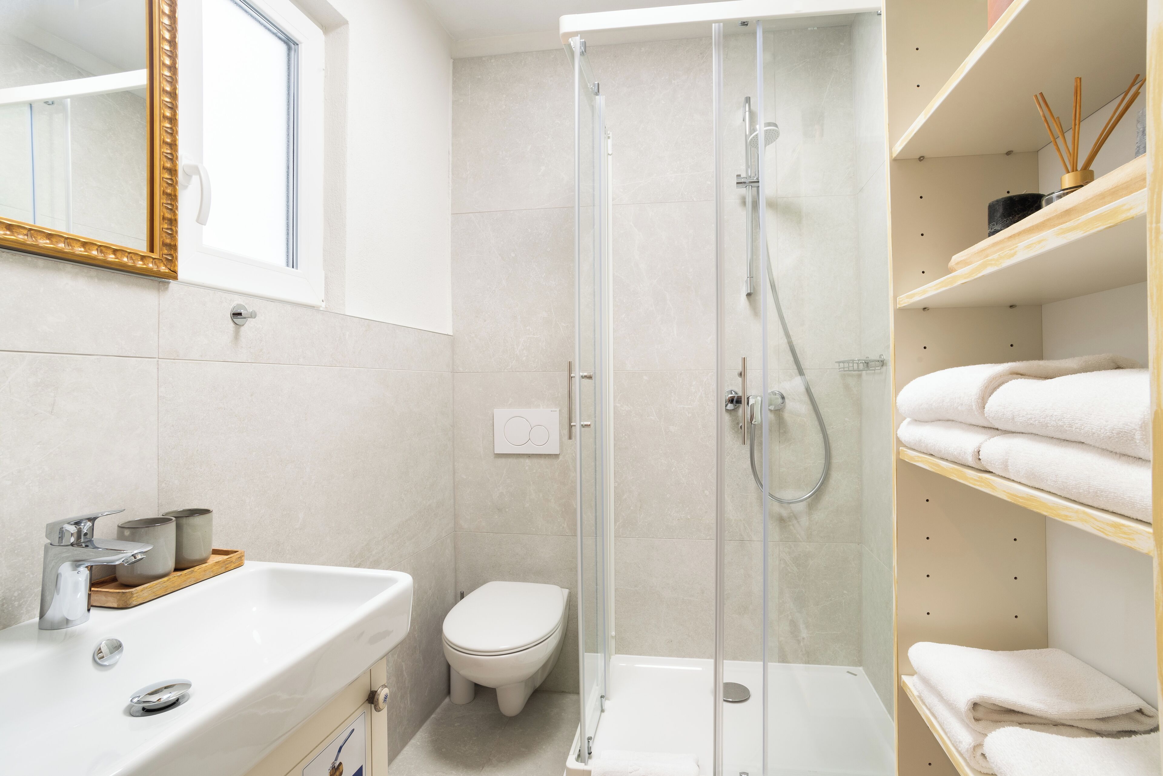 A modern bathroom with a glass-enclosed shower, a white sink, and a toilet. There are shelves with towels and a mirror with a gold frame.