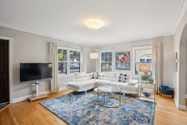 A modern living room with a sectional sofa, a flat-screen TV, a coffee table, and a large window with curtains. - Historic 3BR in Third Ward Walk to UWEC (Eau Claire)