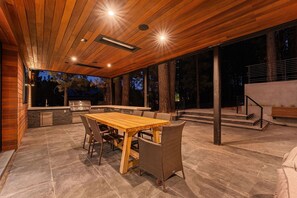 Een terras buiten met een houten tafel en stoelen, een barbecue en een stenen vloer.