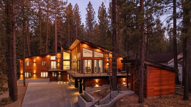 A modern log cabin with a spacious deck and large windows, surrounded by tall pine trees.