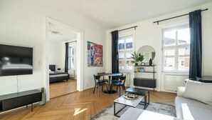 A modern living room with a sofa, coffee table, and a TV mounted on the wall. There is a dining area with a table and chairs, and a large window with curtains. - Rochus Stylish Stays (Vienna)