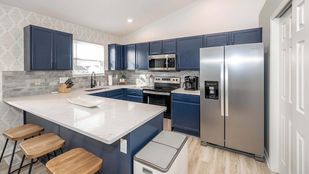 A modern kitchen with blue cabinets, stainless steel appliances, a large island with stools, and a patterned backsplash. - Walton Arkside Oasis (Walton)