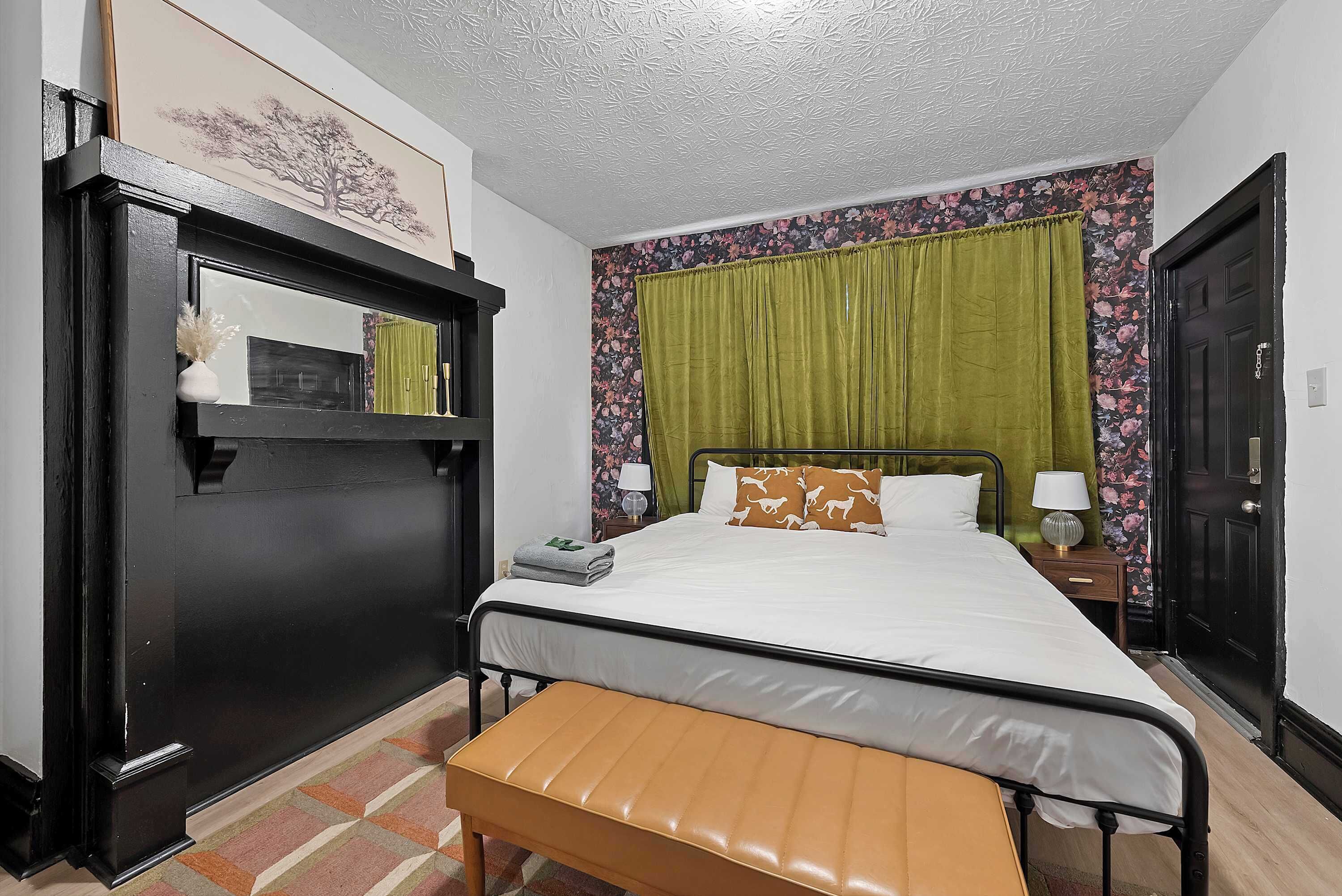 Quarto bem arrumado com cama, um banquinho, mesa de cabeceira, espelho e porta.