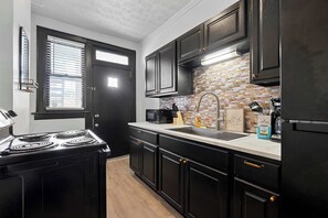 A kitchen with black cabinets, a stove, a sink, and a brick backsplash. - Merion Village Residences 1166 (Columbus)