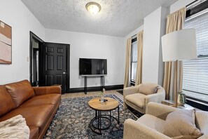 A living room with a brown leather sofa, beige armchairs, a round wooden coffee table, and a flat-screen TV on a stand. - Merion Village Residences 1166 (Columbus)