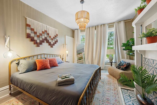 A bedroom with a bed, a hanging light fixture, a window with curtains, and a plant on a shelf.