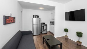 A modern living room with a grey sofa, a black coffee table, a kitchen area with a stainless steel refrigerator, a gas stove, and a microwave. There are two potted plants and a flat-screen TV mounted on the wall. - Northside Suites 3 – A Chic, Relaxed Stay for 2 (Cincinnati)