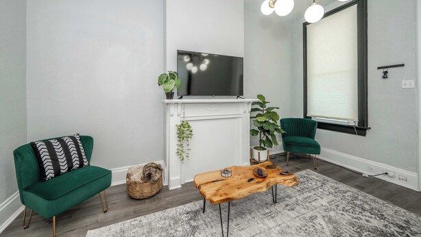 A living room with a green sofa, a wooden coffee table, a TV mounted above a white fireplace, and a large window with blinds. - Chic, Modern Gem with Style (Cincinnati)