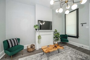Una sala de estar con un sofá verde, una mesa de centro de madera, un televisor sobre la chimenea blanca y un ventanal con persianas.