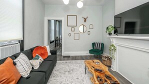 A modern living room with a dark sofa, a wooden coffee table, and a green armchair. There is a fireplace and a flat-screen TV mounted on the wall. - Chic, Modern Gem with Style (Cincinnati)