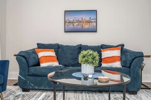 A modern living room with a dark blue sofa, two orange and white striped pillows, a glass coffee table with a blue coaster, and a framed cityscape picture on the wall.