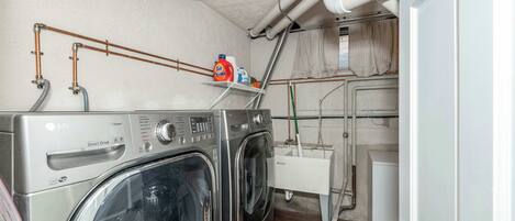 A laundry room with two stacked washers and dryers, a shelf with cleaning supplies, and visible plumbing.